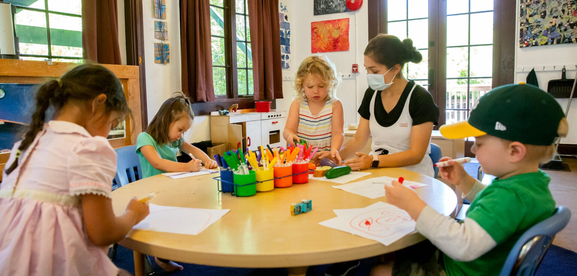 A childcare worker and four children are gathered around a table and drawing on paper.