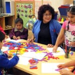 An early educator smiling at a student while a group of students are learning how to draw letters