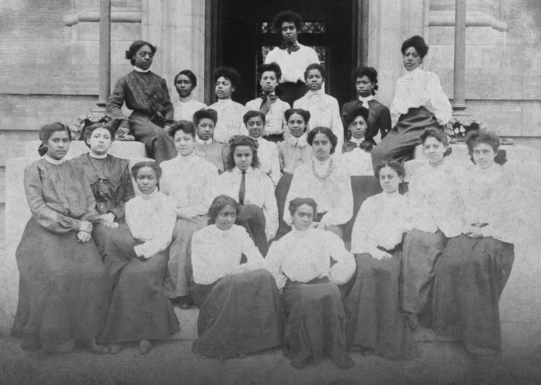 A group of Black women are seated outside of a building and posing for a formal black and white portrait.