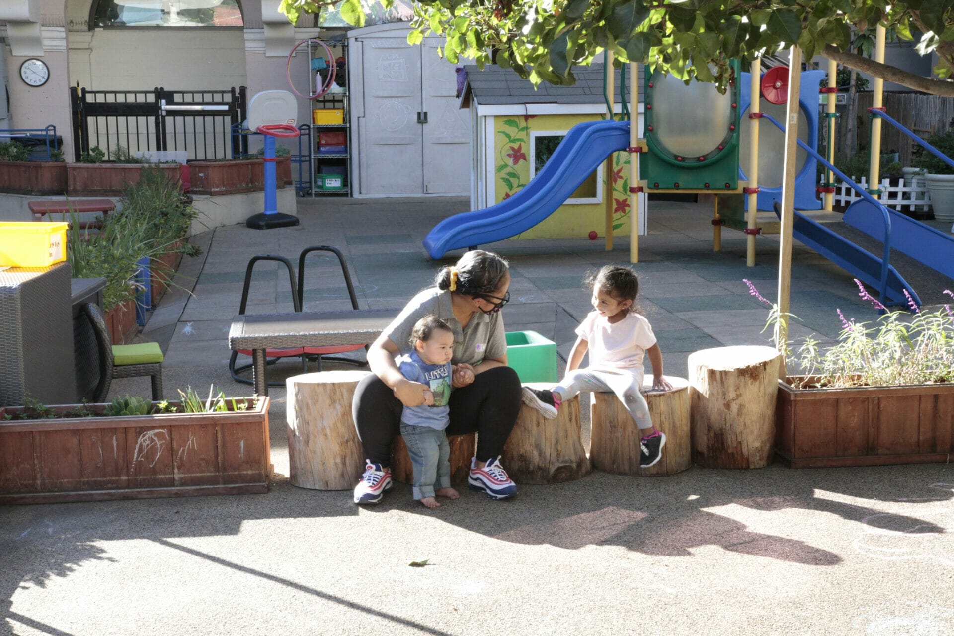 A child educator is sitting outside holding a young child and talking to another child.