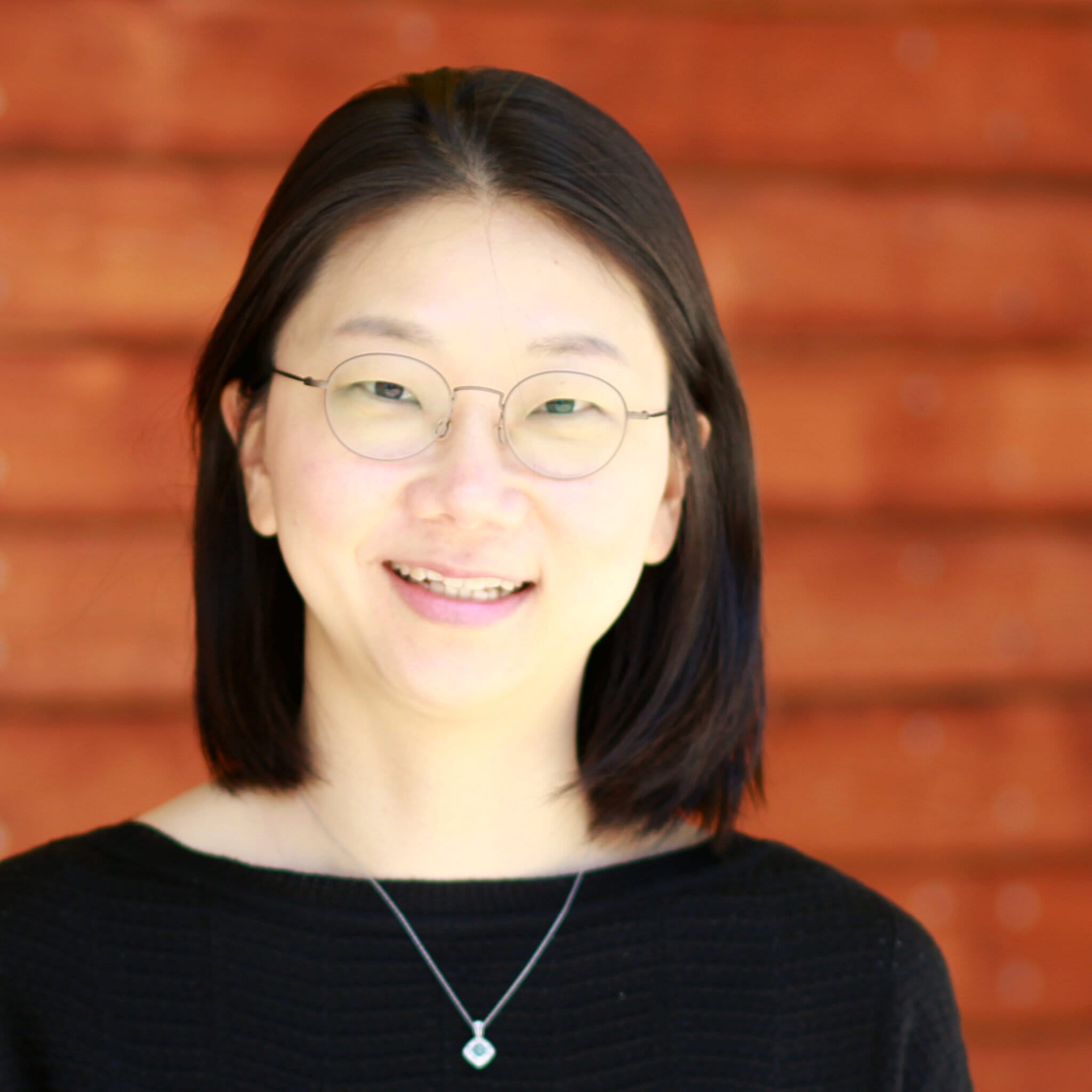 A smiling woman in glasses stands in front of a wooden wall., Yoonjeon Kim