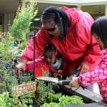 An early educator teaches two children how to use the gardening tools for the outdoor garden.