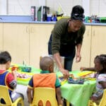 An early educator is helping children at a table with the paintbrushes.