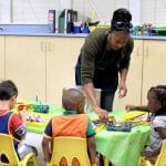 An early educator is helping children at a table with the paintbrushes.