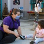 Masked early educator is laughing with a child while sitting outside.