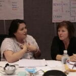 Two women are speaking to each other at a table with notes laid out in from of them