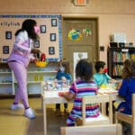 A masked educator stands next to a table where children are playing.