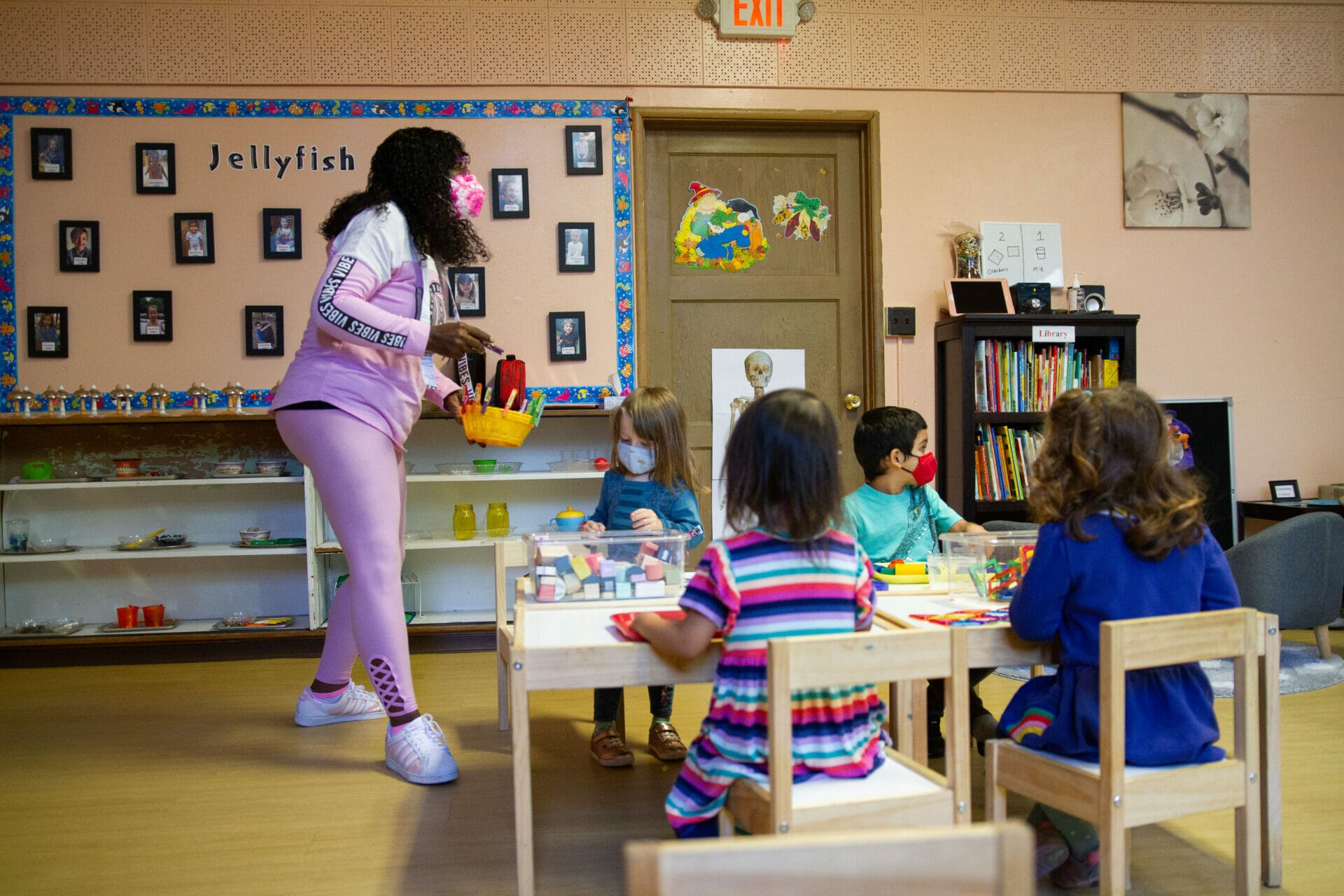 A masked educator stands next to a table where children are playing.