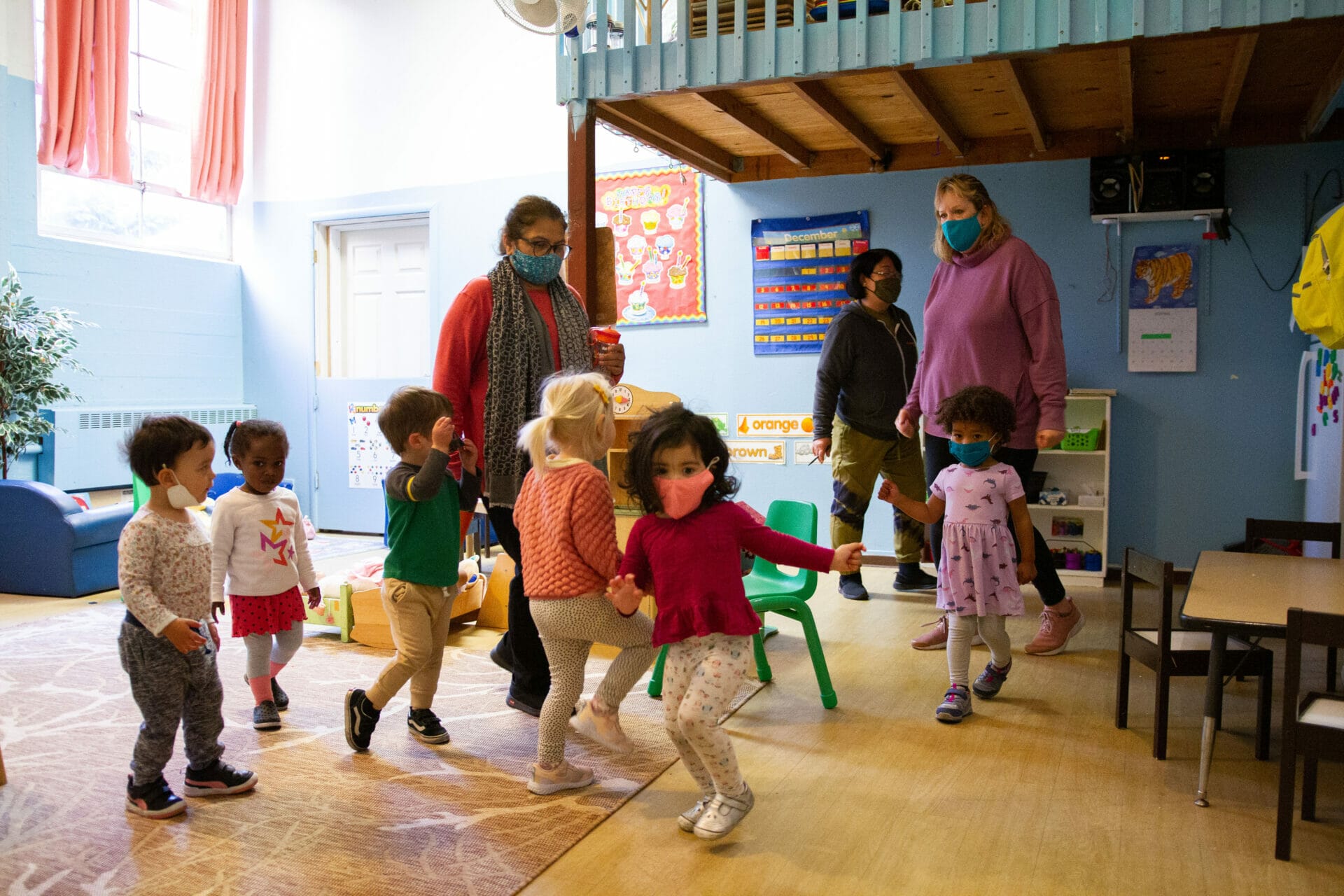 A group of masked children and three masked early educators are dancing inside a classroom.