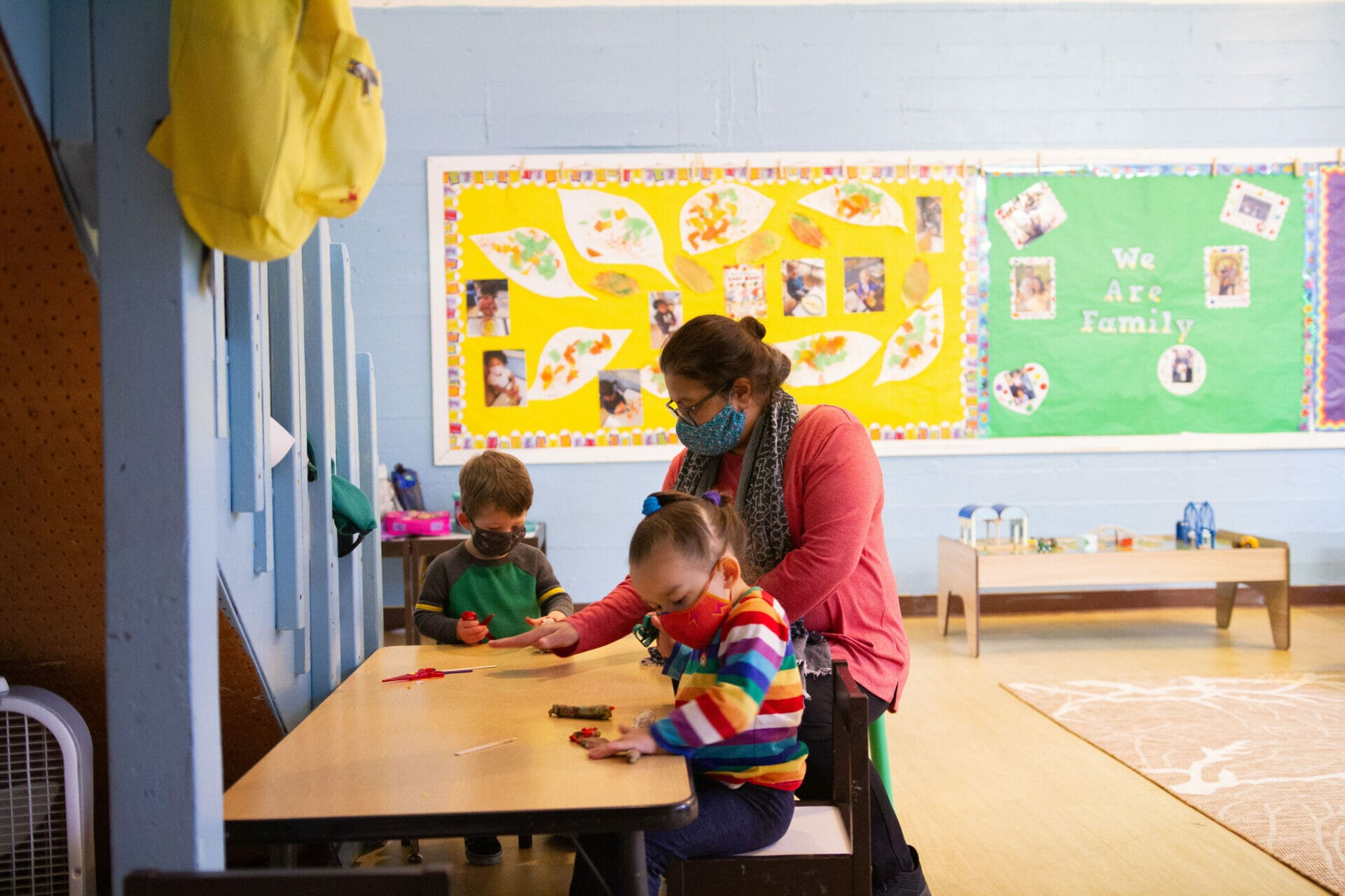 Masked early educator assisting two students on an activity inside a classroom