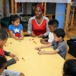 An early educator is explaining an block activity to a group of children at a table.