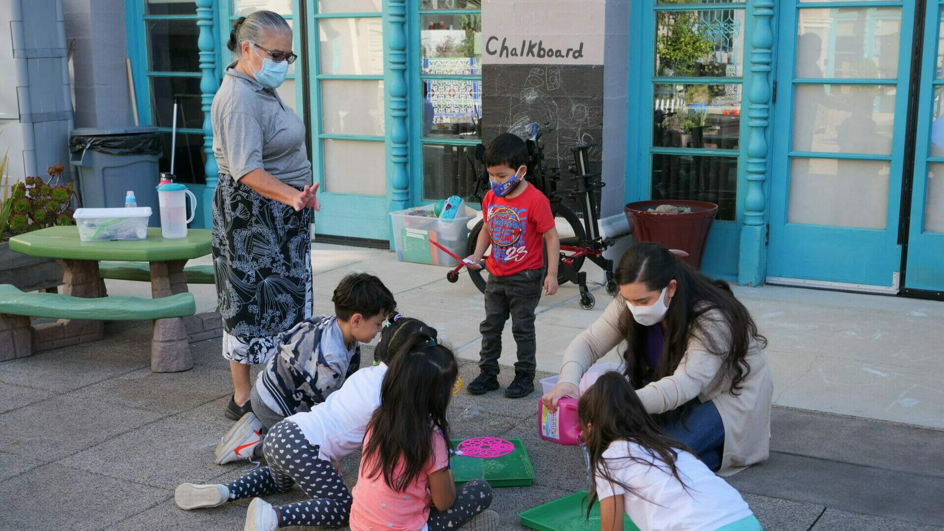 A group of young children gather around a crouching early educator who is making bubbles while another teacher in a surgical mask stands next to them.