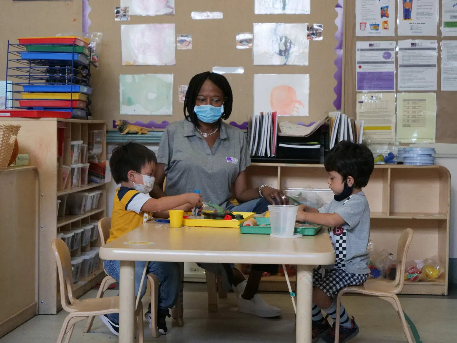 A Black early educator wearing a surgical mask sits with two young children playing with dinosaur toys and sand at a children's table in a classroom.