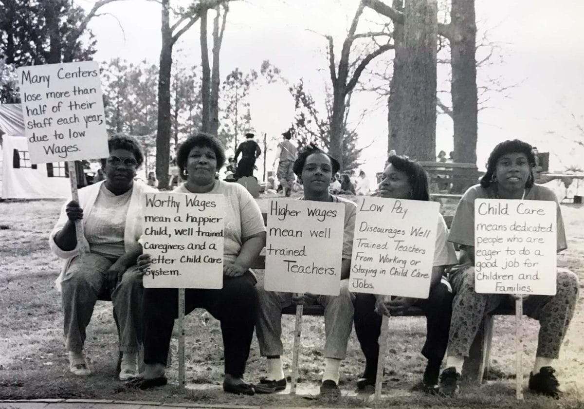 Early educators seated in a park holding protest signs with slogans like, "Worthy Wages mean a happier child, well-trained teachers, and a better childcare system."