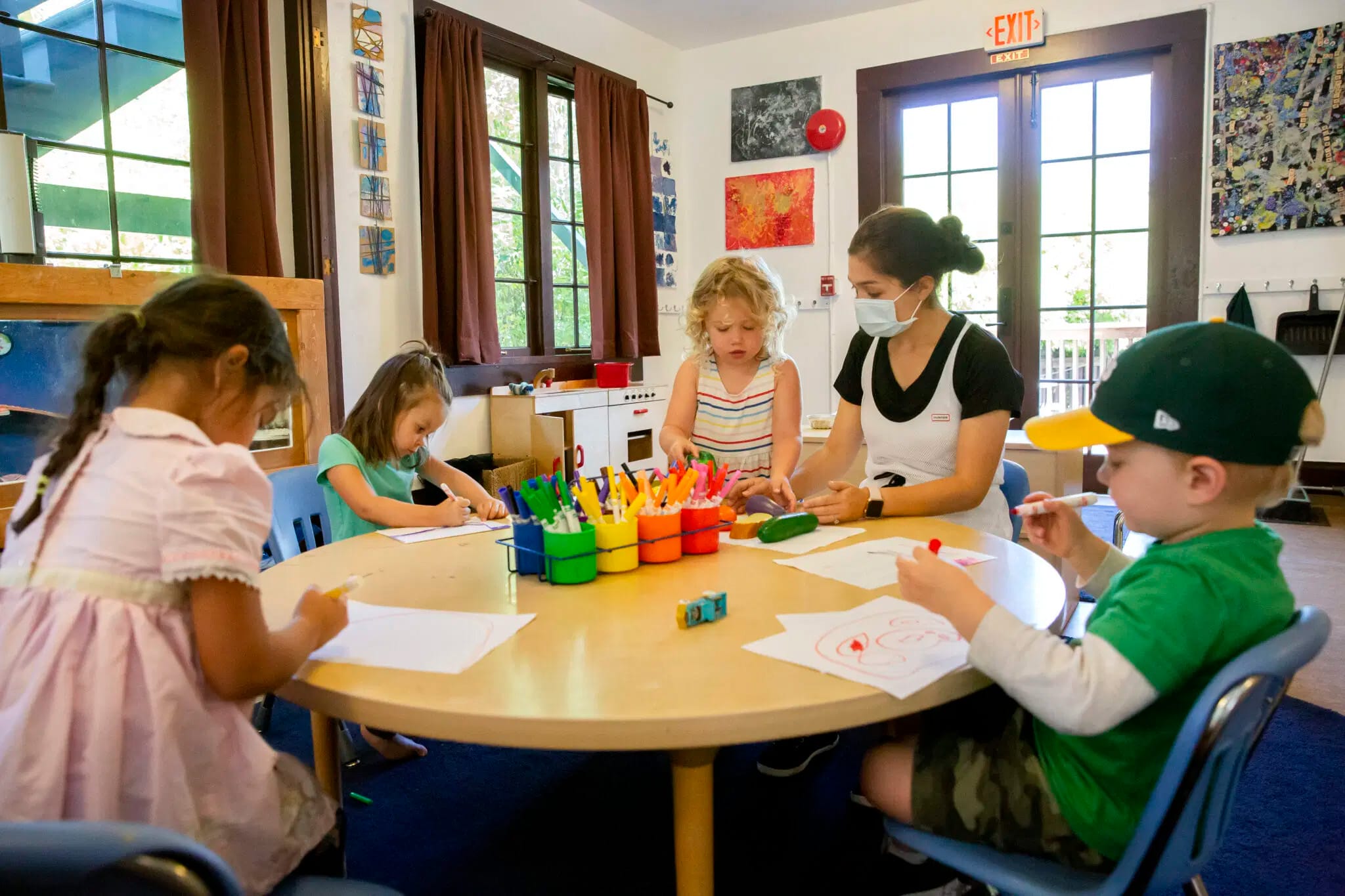 A Child Care Teacher With Four Children Around A Table Working On Drawings