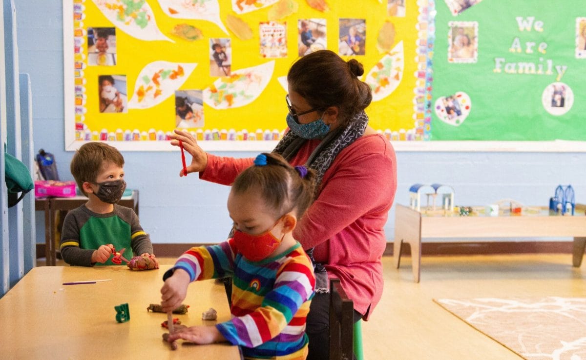 A masked educator, a young boy and a young girl sit at a classroom table while playing with clay.