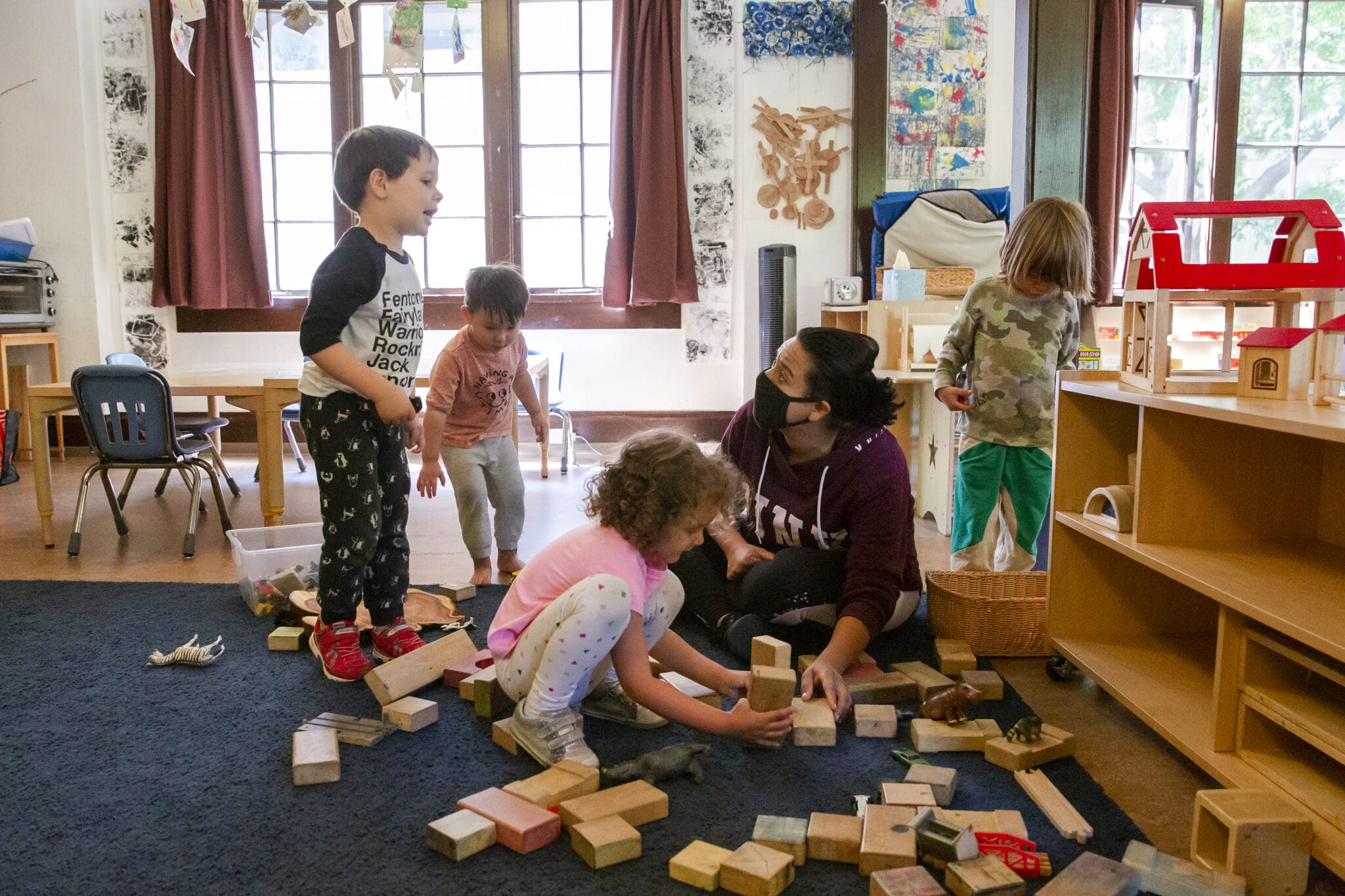 A teacher pictured sitting in the block area of a classroom talking to a child on their left while supporting another child with block play.