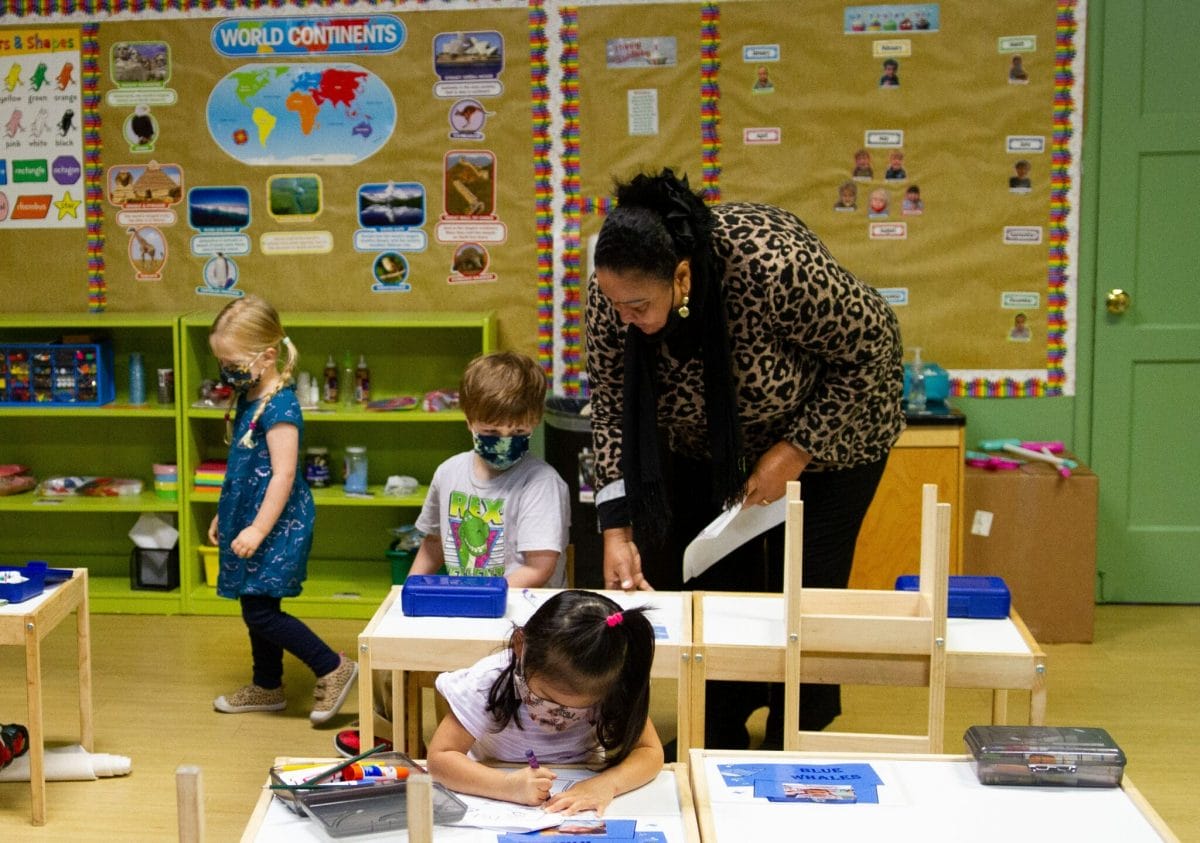 Two early educators sit next to young students in a bright green, well-lit classroom. The educators assist the children with writing in their notebooks.