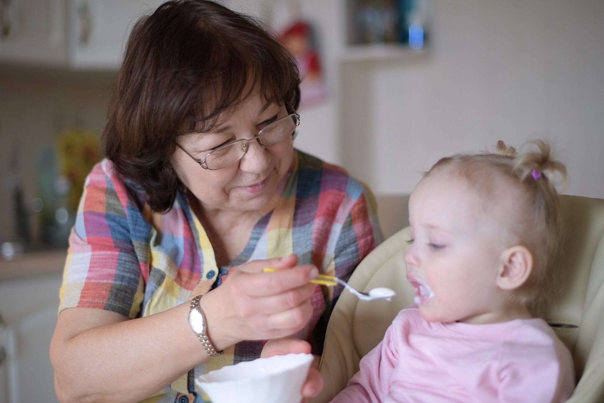 A woman in her 60s feeds a baby in a high chair.