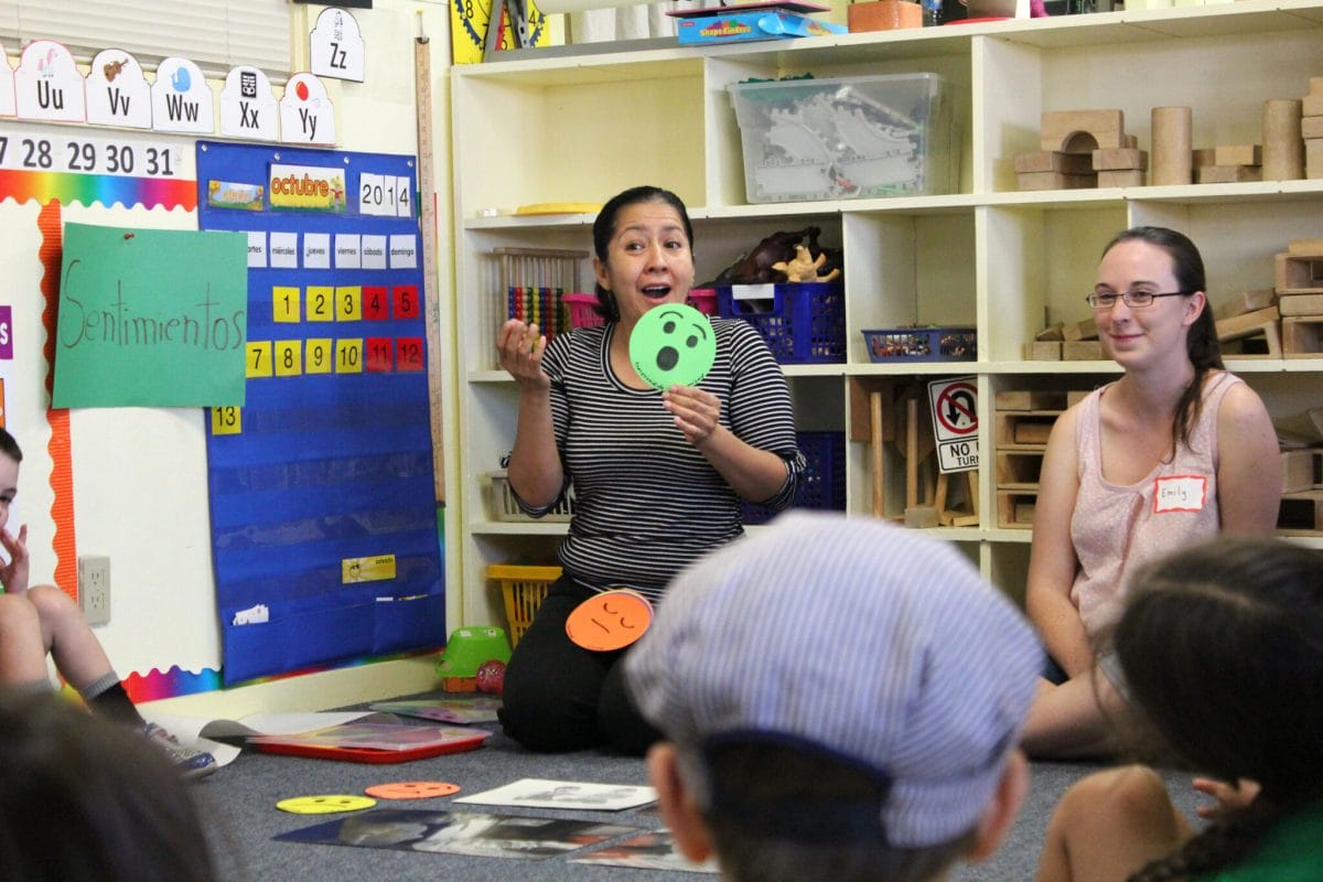 An early educator surrounded by children holds up a green smiley face cue card with a joyful expression. A sign behind her says “sentimientos."