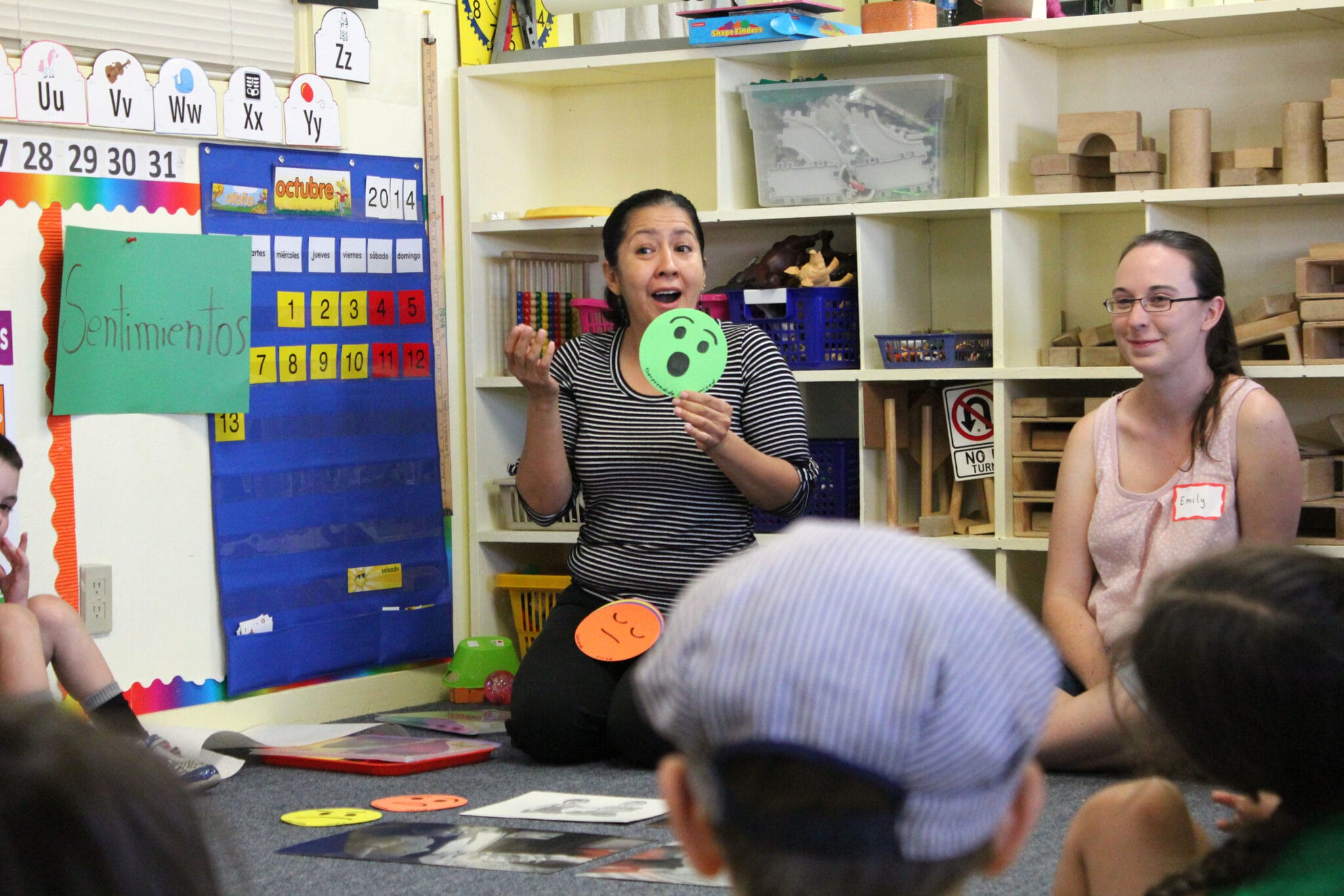 An early educator surrounded by children holds up a green smiley face cue card with a joyful expression. A sign behind her says “sentimientos.