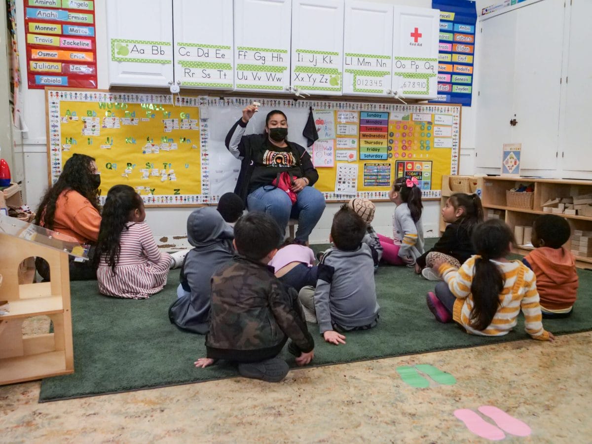 A teacher sits with her circle of young children in a classroom decorated with letters and numbers.