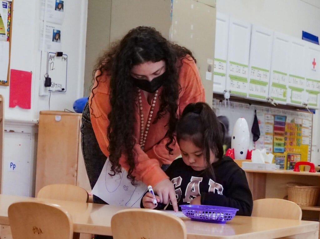 An early educator wearing a mask bends over a desk and talks to a toddler who is drawing a picture.