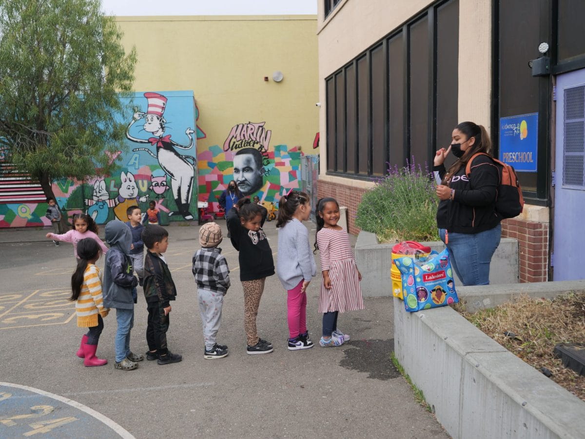 A teacher wearing a backpack gives instructions to her preschool students as they line up to go inside.
