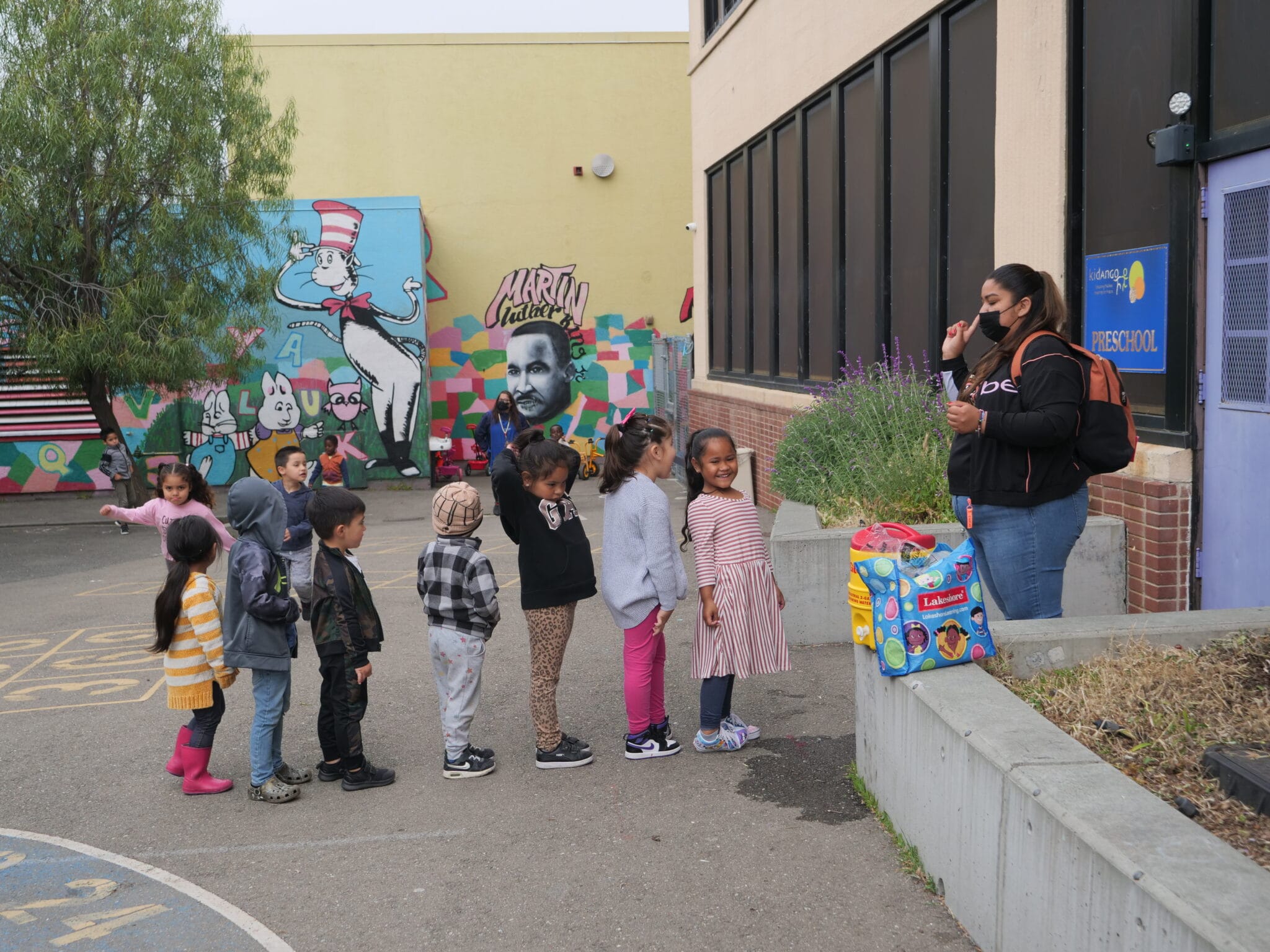 A teacher wearing a backpack gives instructions to her preschool students as they line up to go inside.