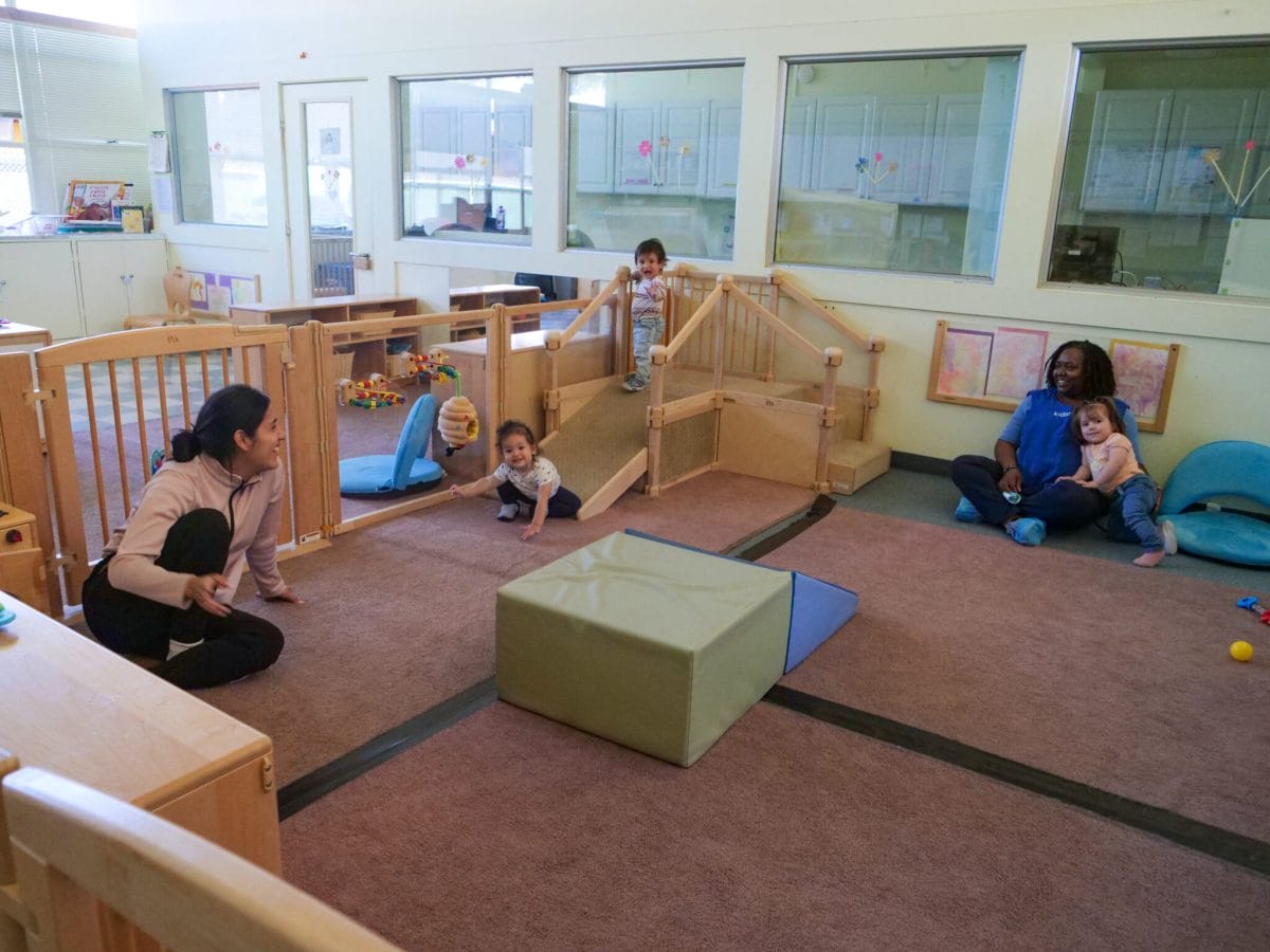 Two educators are sitting apart from each other in an indoor play area with three toddlers playing alongside them and smiling at the camera.