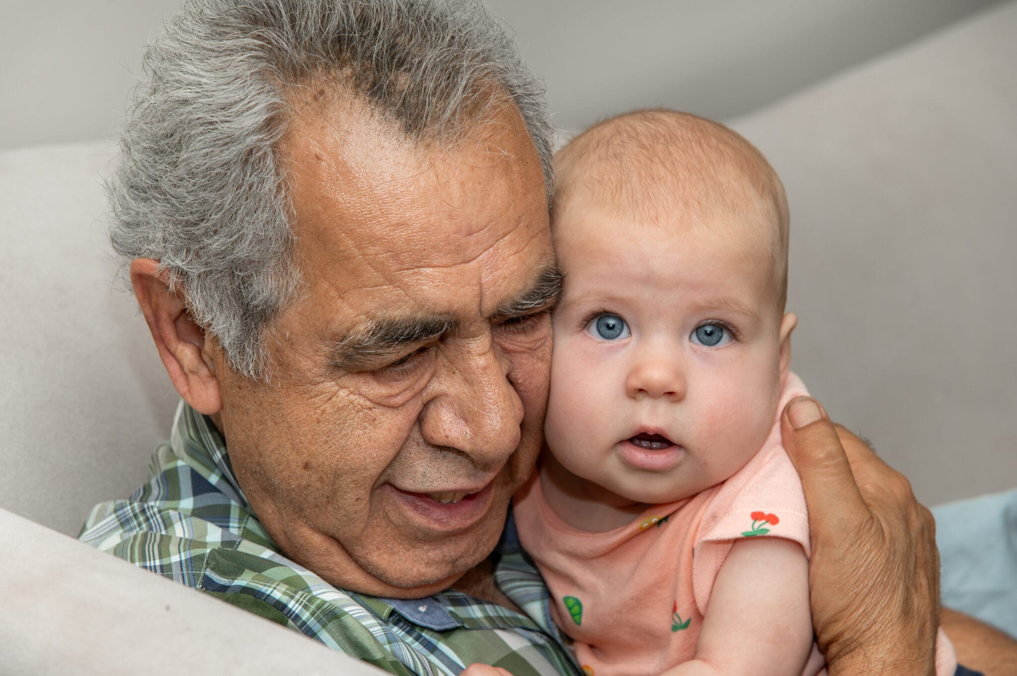 A wrinkled old man with short, silver hair cuddles an infant gently up to his cheek.