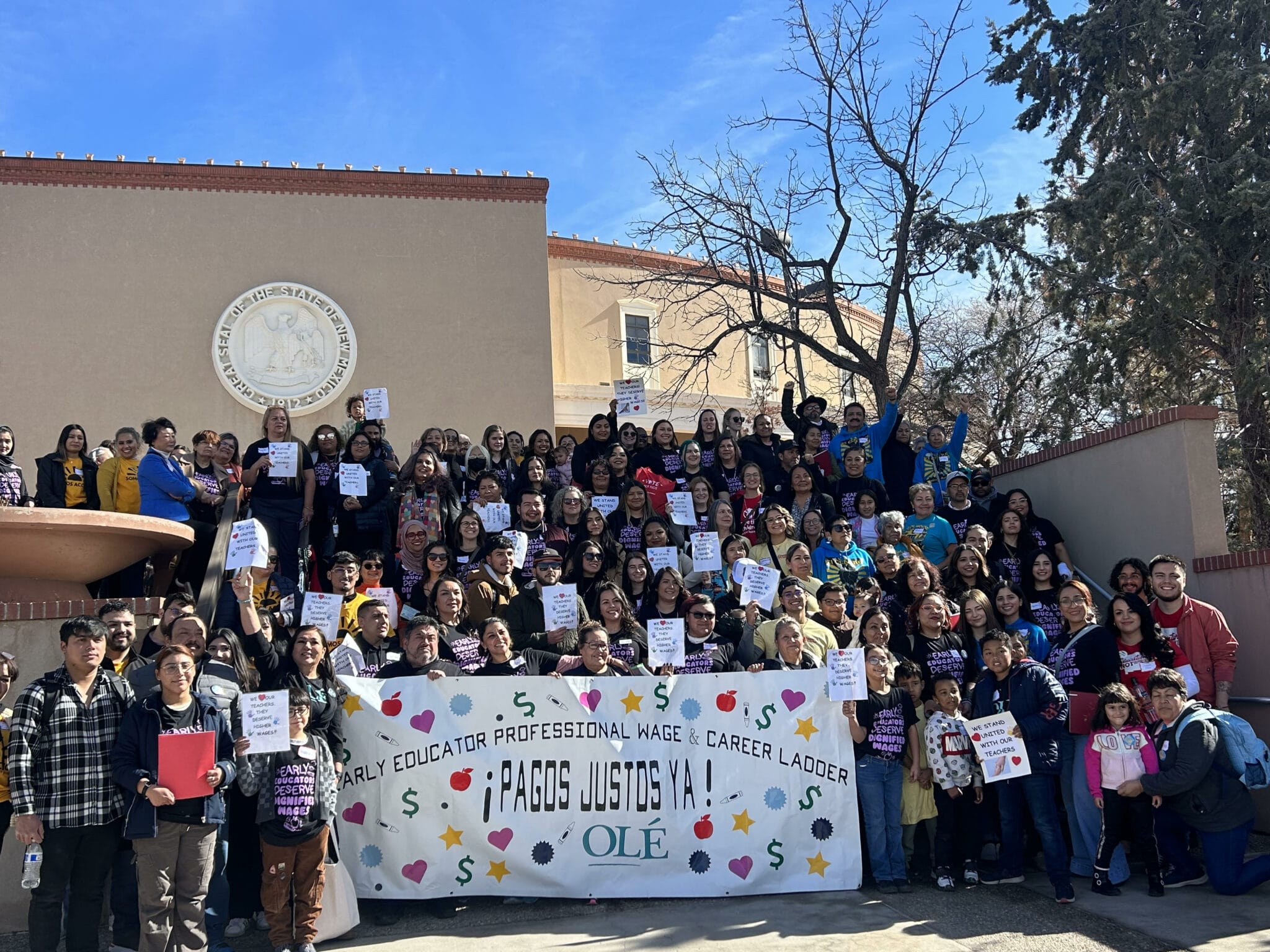 A large group of educators, children and community members gather on stairs behind a sign that reads “Early Educator Professional Wage and Career Ladder, Pagos Justos Ya!”