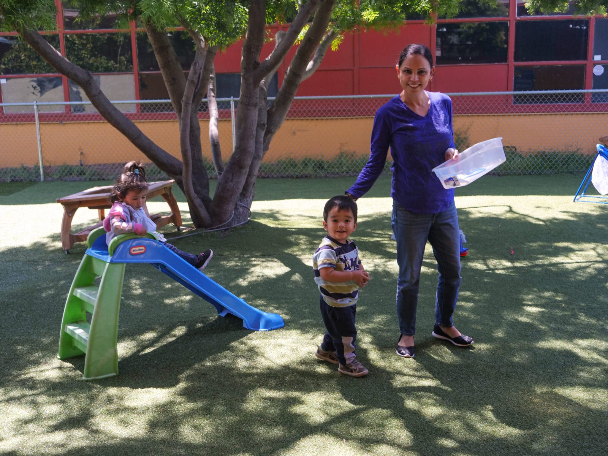 An educator pauses to smile at the camera during outdoor play with two toddlers.