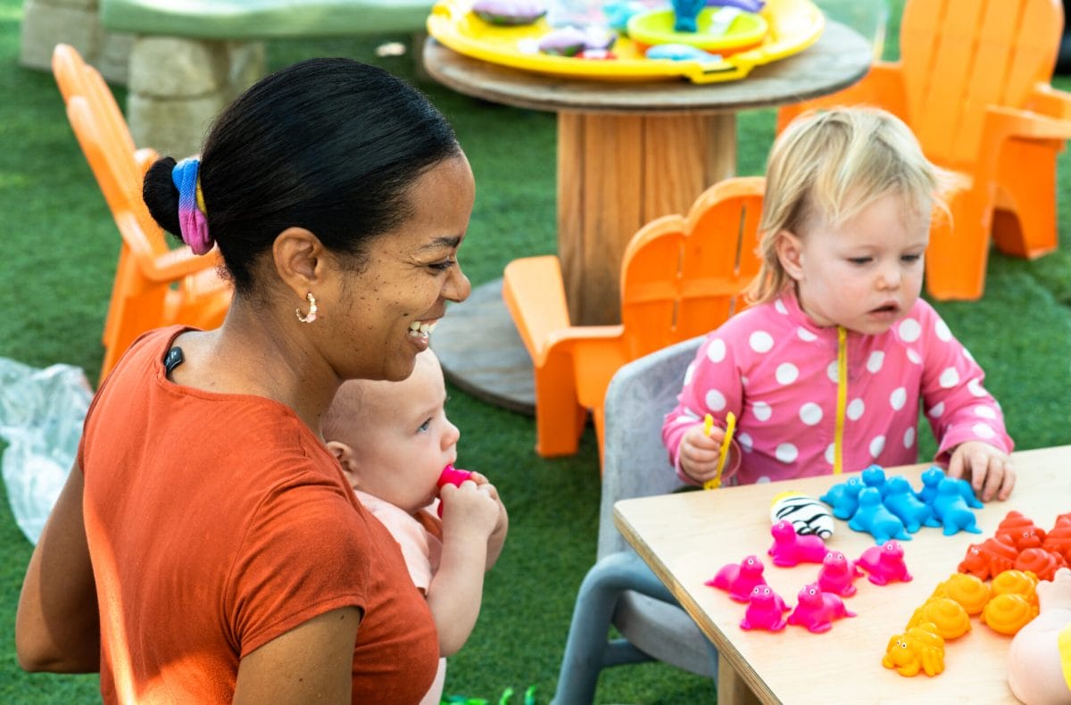 A smiling educator holds a baby and kneels next to a toddler playing at a table.