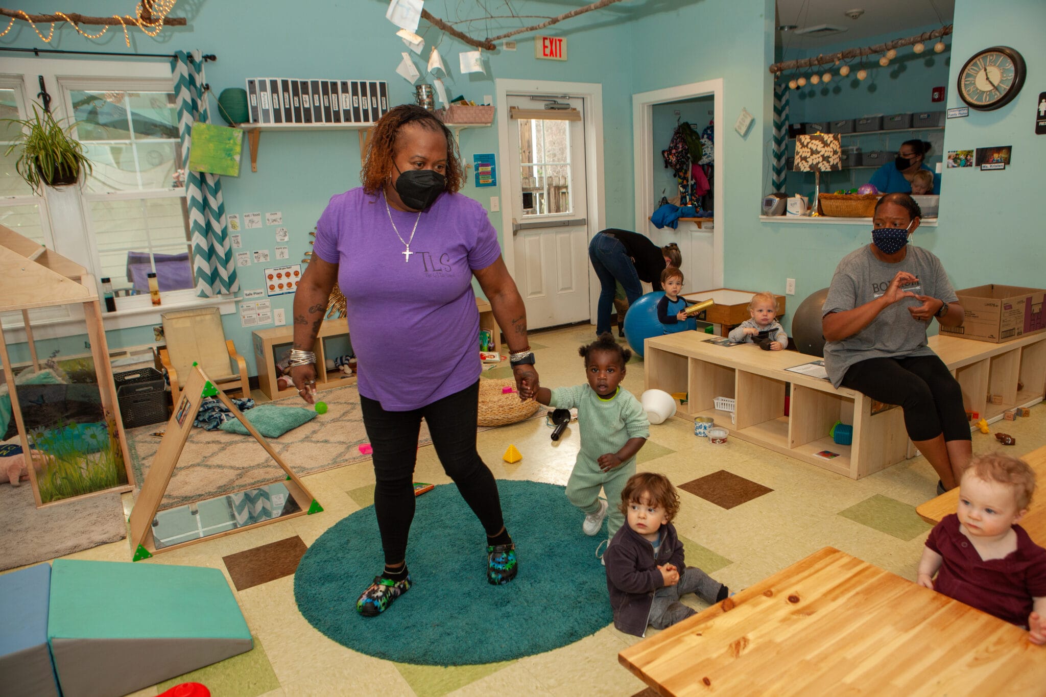 An early educator in a purple shirt holds a toddler’s hand in the middle of a bright classroom surrounded by four other infants. Another early educator sits making hand motions to a child, while a third educator bends over to pick something up.