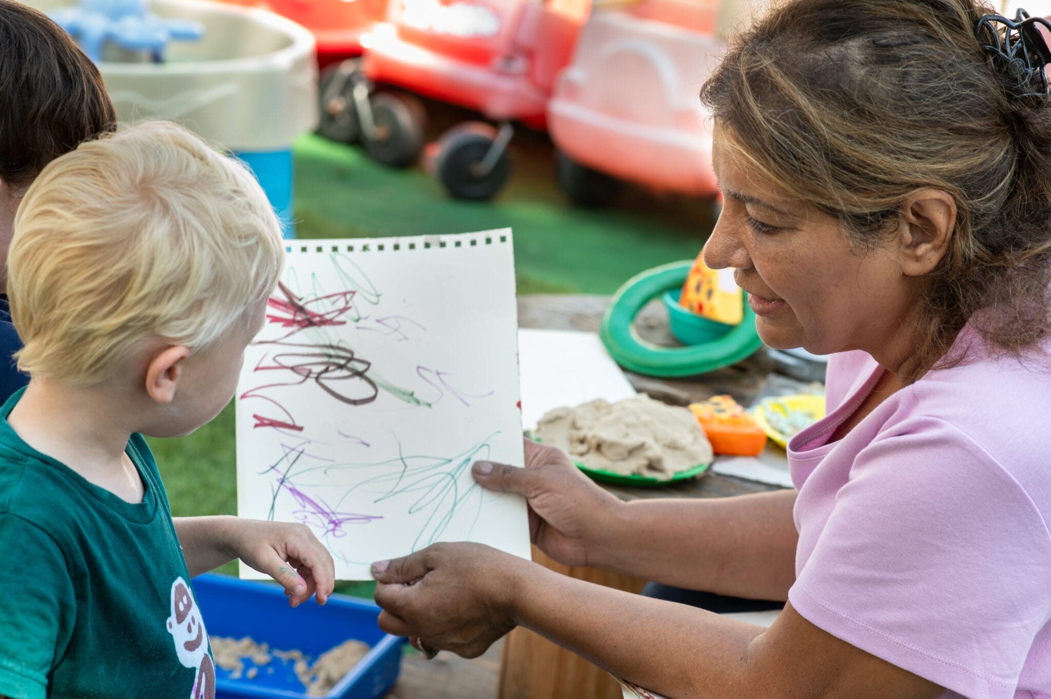 A woman in a pink shirt holds up a child's colorful drawing as they share a moment of creativity outdoors.