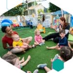 Two smiling educators sit in a circle on astroturf with a group of young children