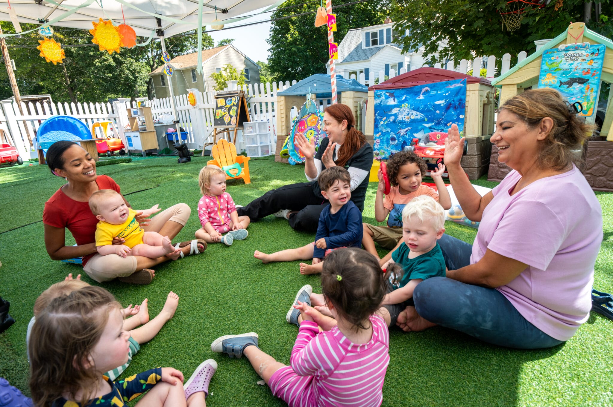 Child care workers and children sitting in a circle outside in a play area.