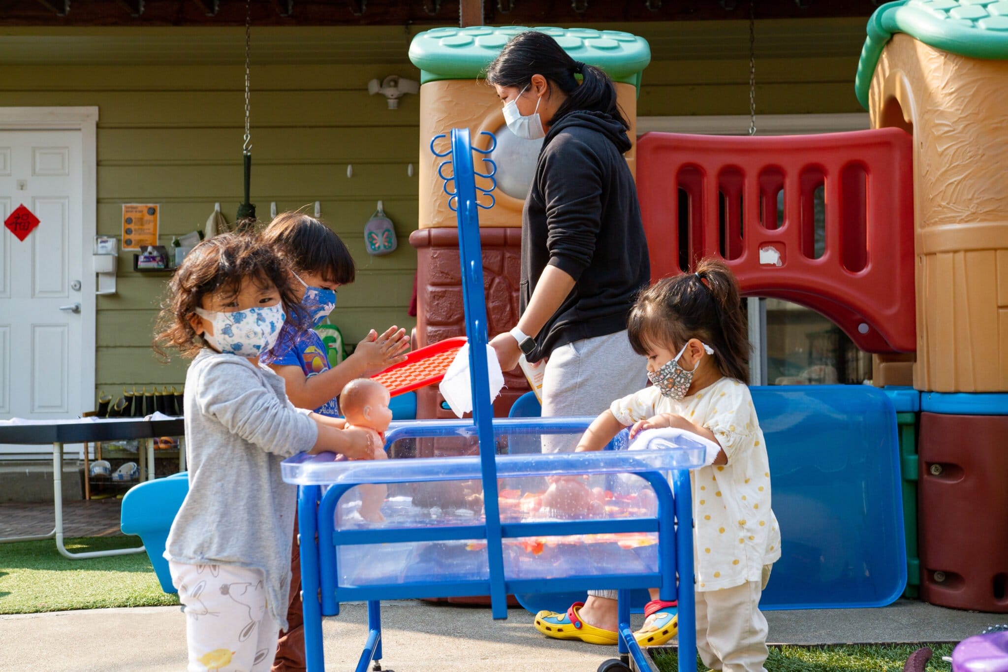 Three masked children play outside while a mask caregiver sanitizes equipment behind them.