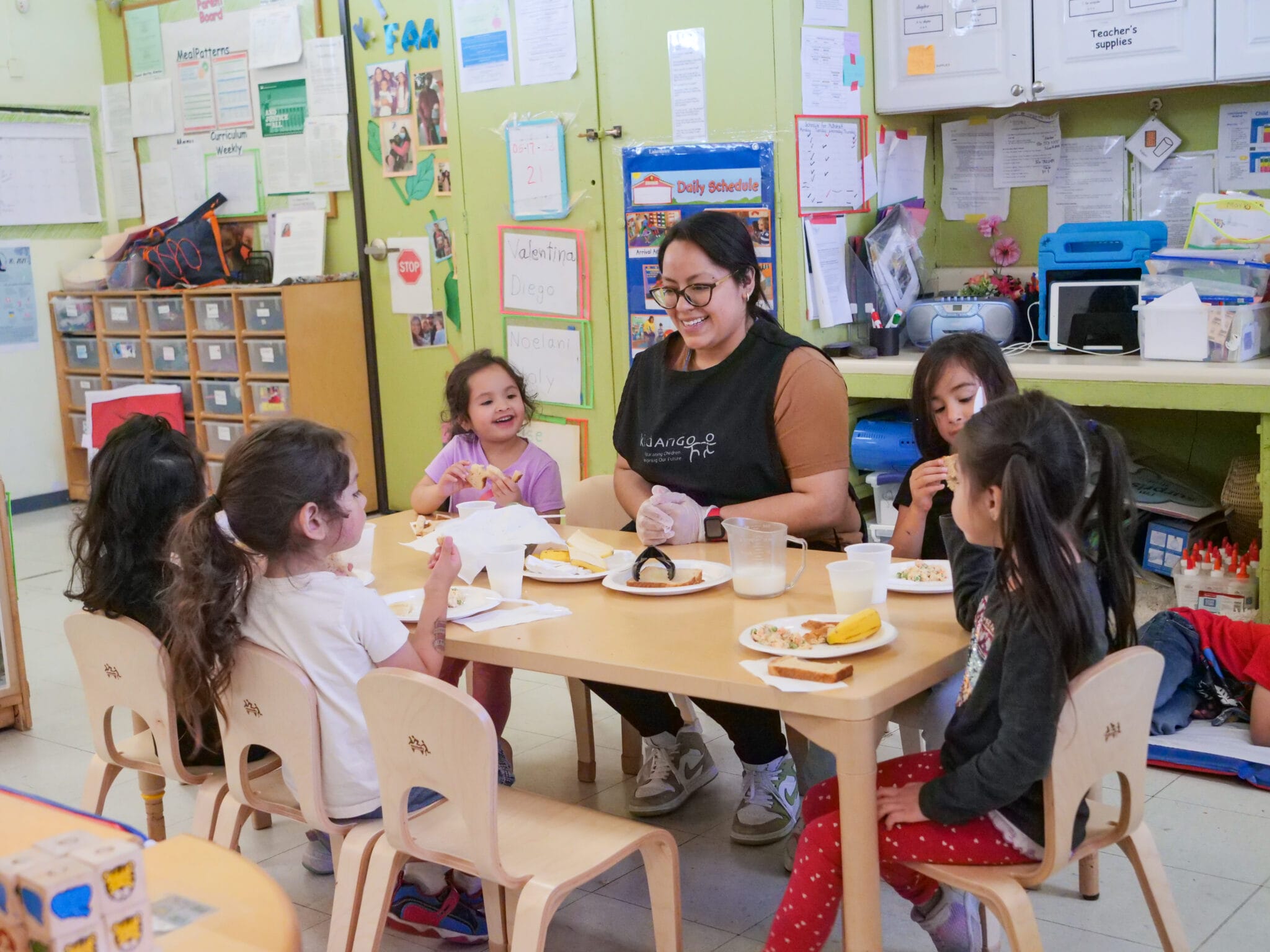 A teacher sits at a table with five children while they eat their lunch.
