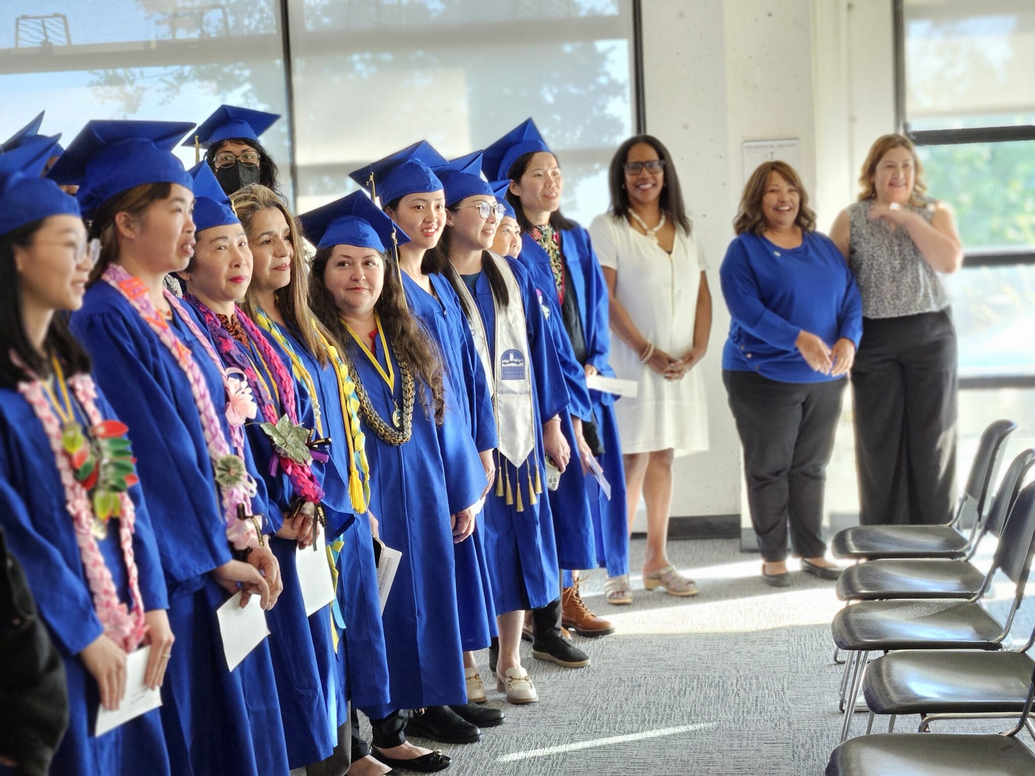 Graduates in blue caps and gowns stand together during a ceremony with faculty and staff looking on.
