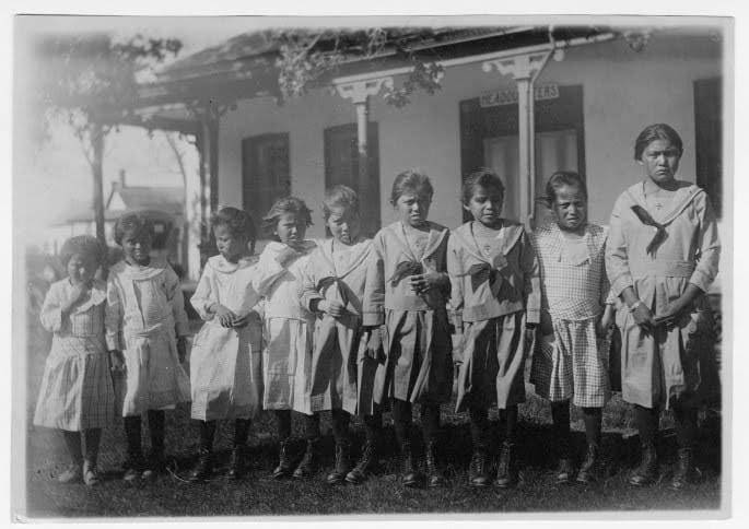 A black-and-white archival photograph from 1923 showing ten young Indigenous girls standing in a line in front of a school building, all wearing matching western-style dresses.
