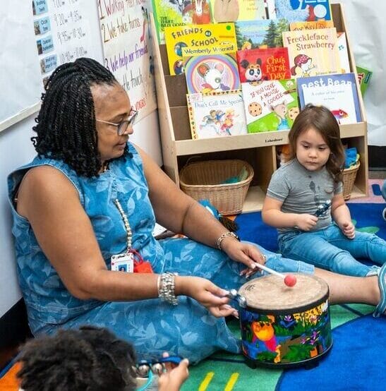 A teacher and students sit in a circle on the classroom floor.