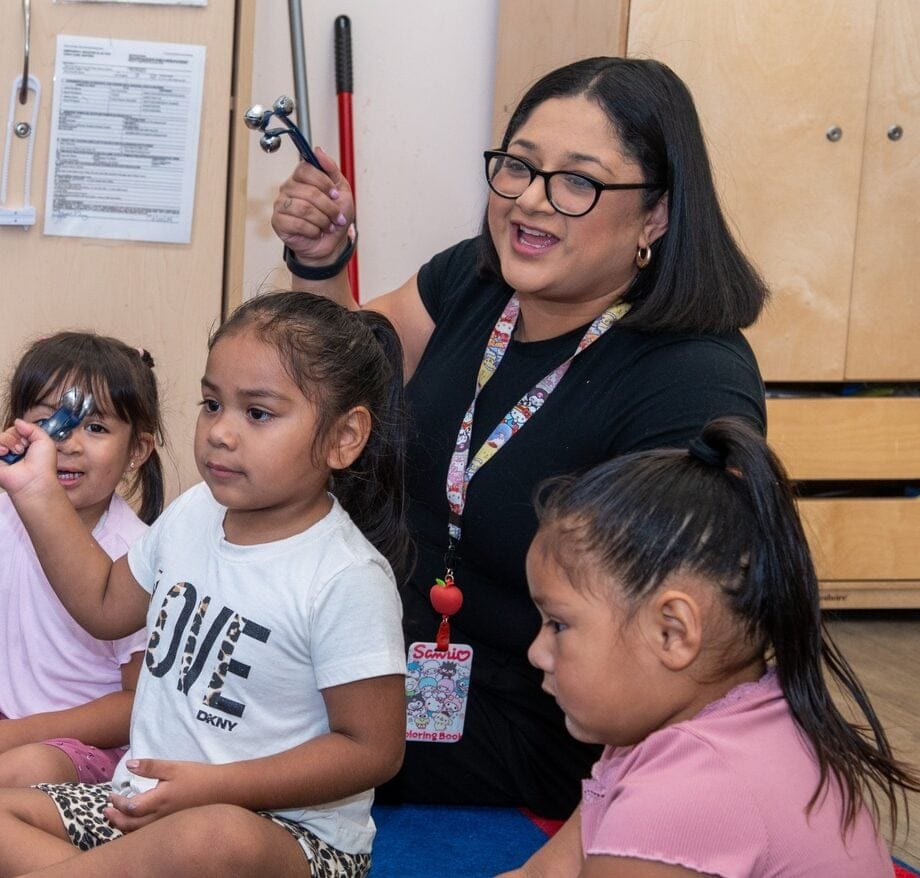 A teacher and students sit in a circle on the classroom floor.