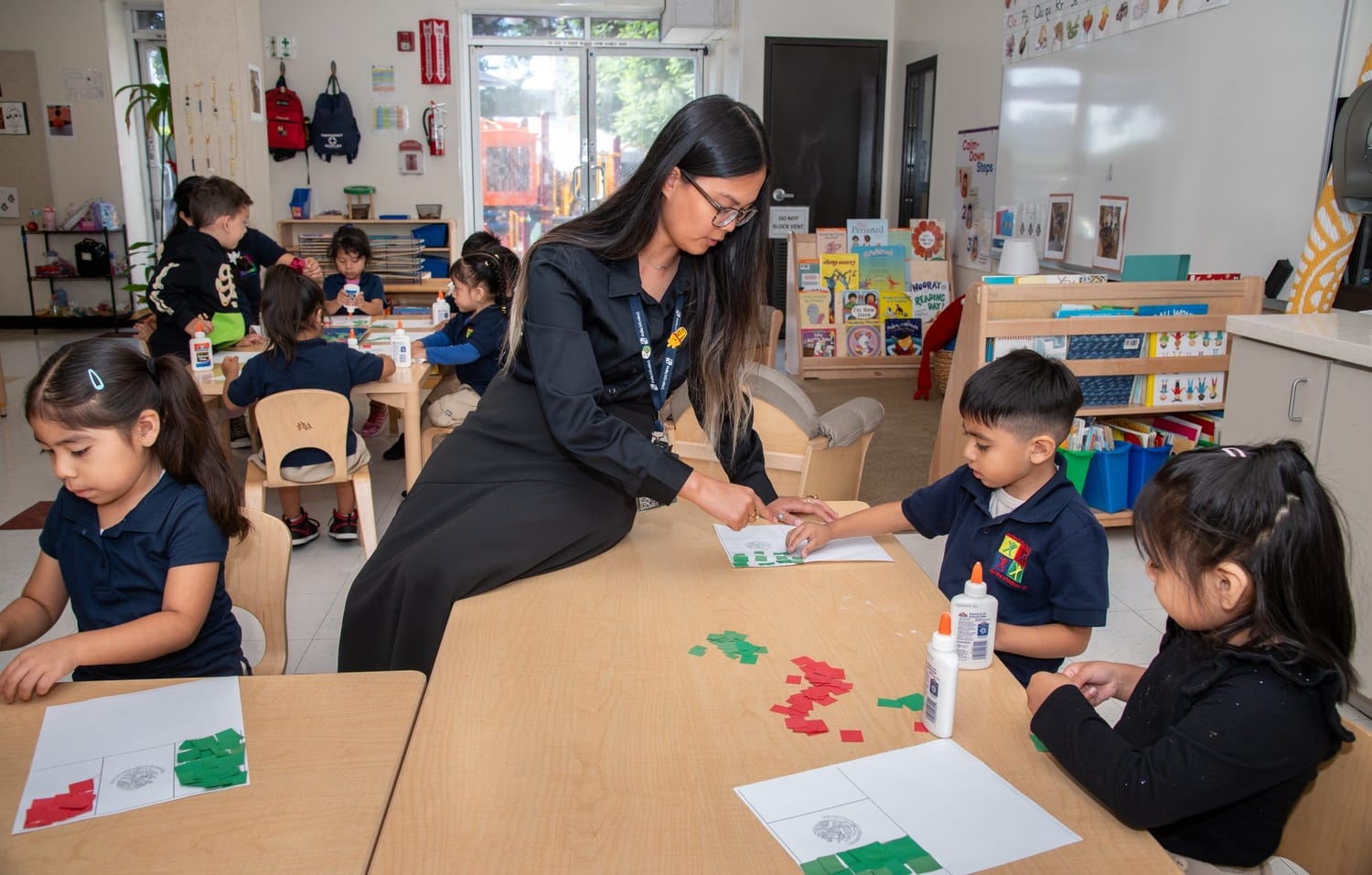 A teacher leans on a table while helping a student with arts and crafts.
