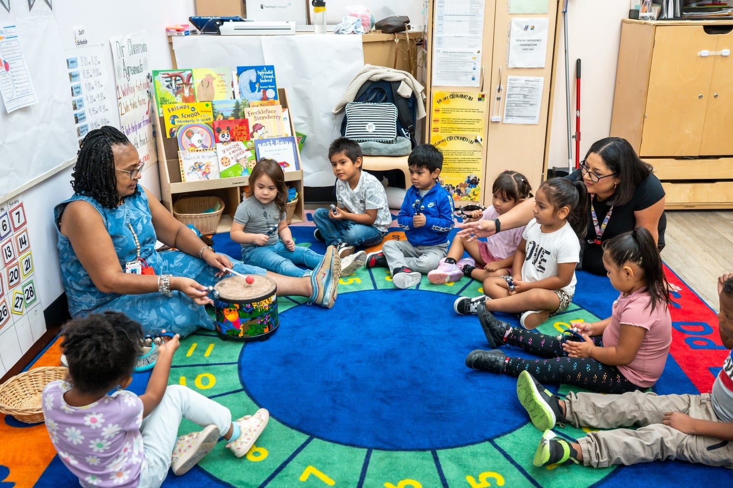 Early childhood educator plays with toy drums for a group of young students holding various instruments.